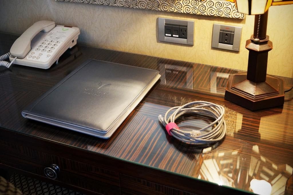 Quarto de hotel com uma secretária de madeira, um laptop, um telefone e uma lâmpada.