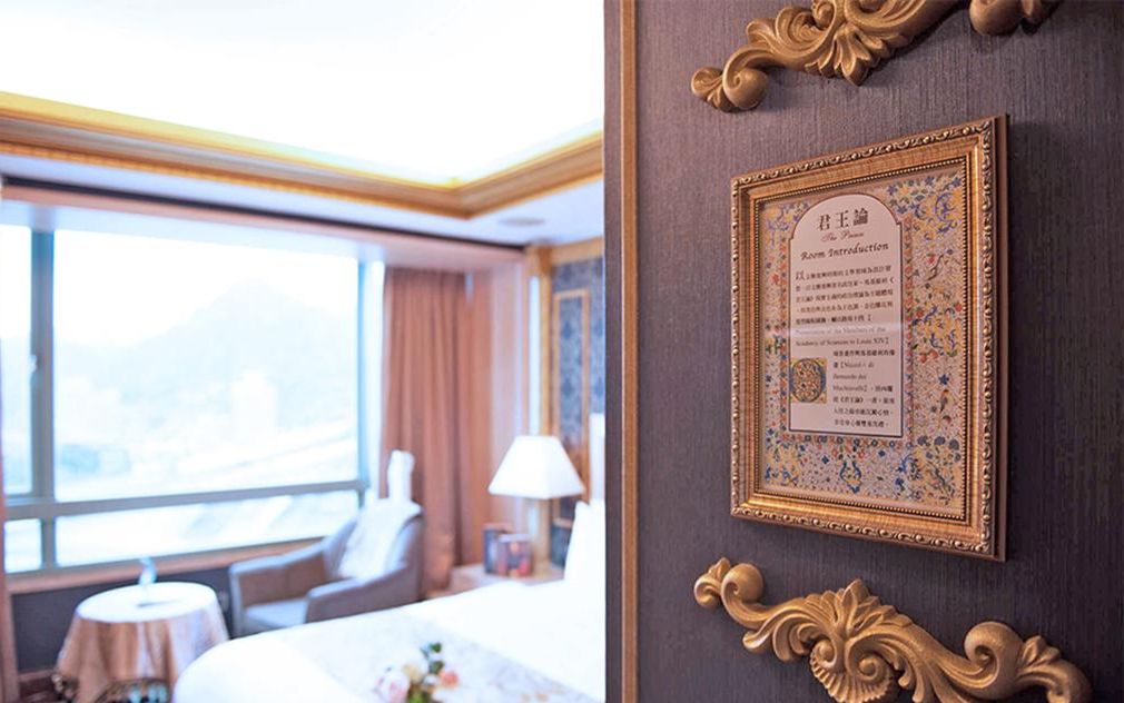 Quarto de hotel com uma janela grande, uma cama, uma cadeira e um certificado emoldurado na parede.