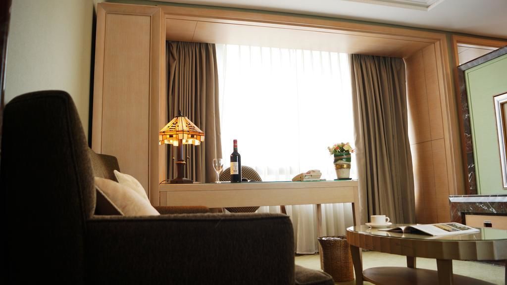 A hotel room with a sofa, a small table with a lamp, a wine bottle, and a view of the outside through large windows.