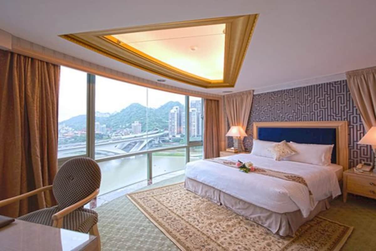 Habitación de hotel con una cama grande, un escritorio y una silla. Se ve un paisaje urbano y un puente a través de la ventana.