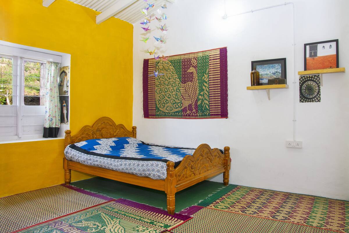 Una habitación con una cama de madera, alfombras coloridas, un ventanal con cortinas y adornos en la pared, incluyendo un tapiz y fotografías enmarcadas.