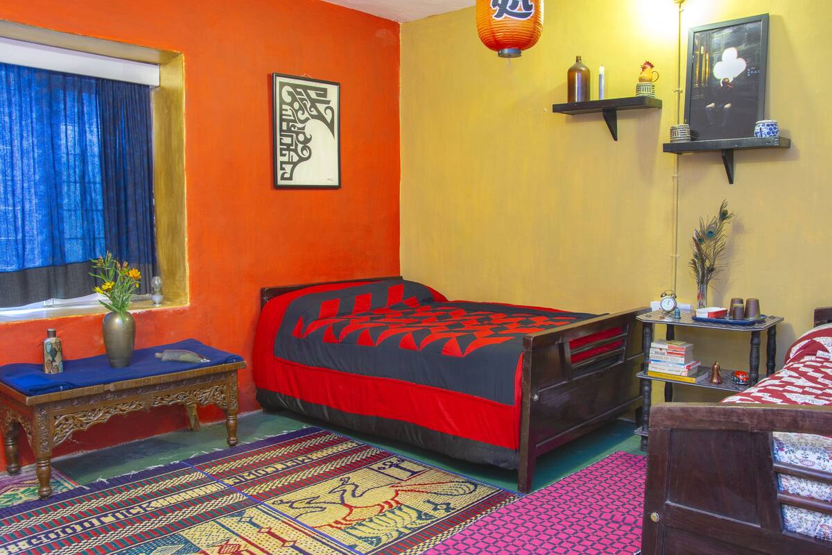 Una habitación con una cama, una ventana con cortinas, una mesa con un florero y una estantería con objetos decorativos.