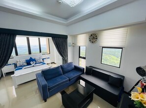 A modern living room with a sofa, a coffee table, and a black couch. There are large windows with curtains, a clock on the wall, and a view of the outdoors. - Splendido Tower 2 Ann's Unit  (Laurel)