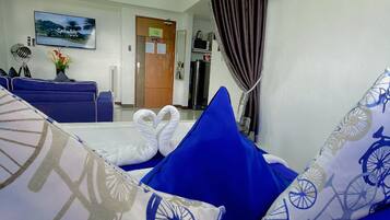 Habitación de hotel con sofá azul, televisor de pantalla plana, cama con toallas blancas y vista al pasillo.