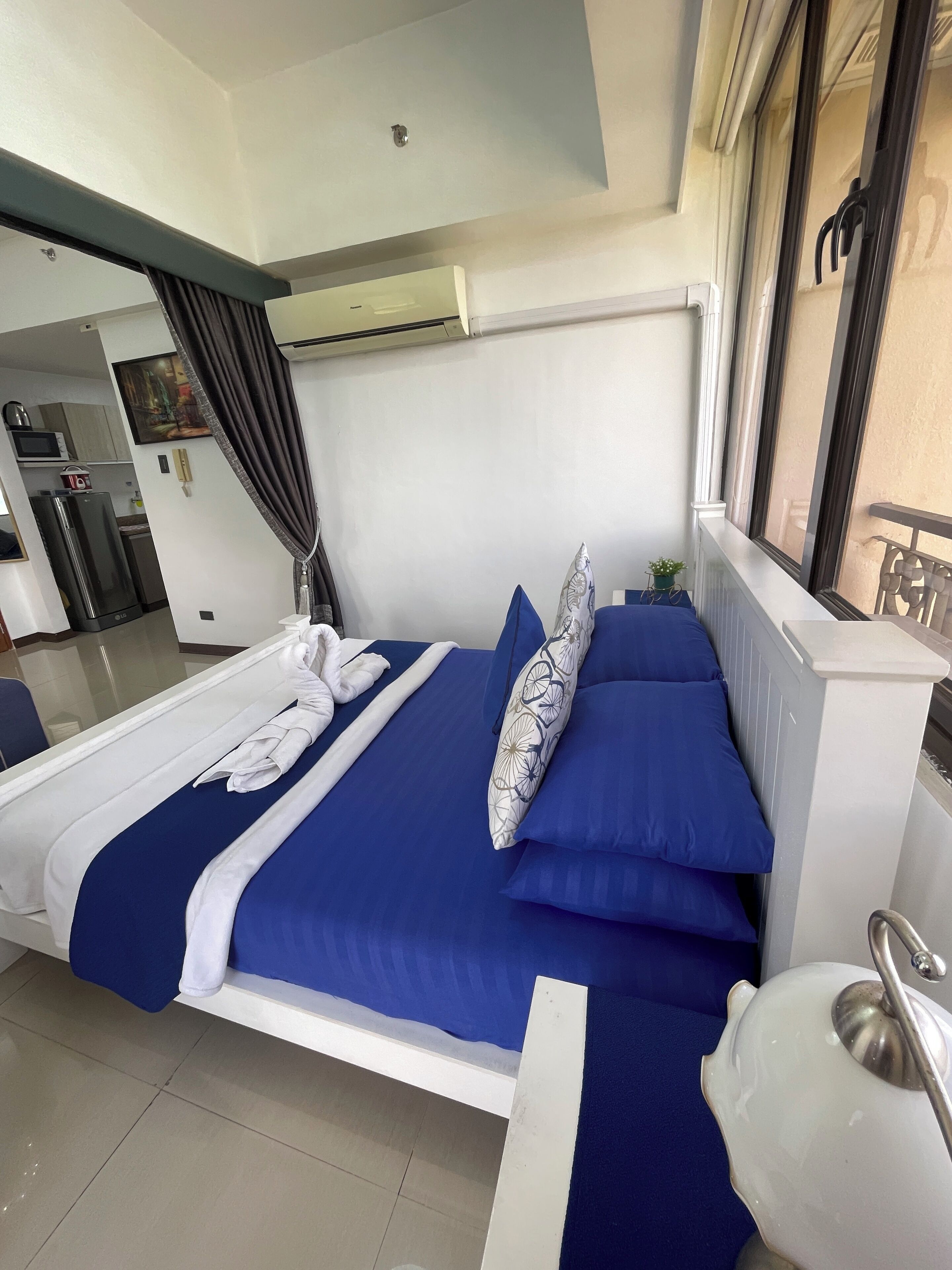 A hotel room with a large bed, a small table, a chair, and a view of the kitchen and balcony.