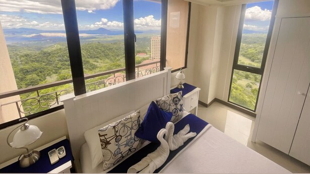 A bedroom with a bed, a nightstand, a lamp, and a view of a green landscape through large windows. - Splendido Tower 2 Ann's Unit  (Laurel)