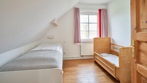 Ein kleiner, gut beleuchteter Raum mit einem Einzelbett, einer Holzbank und einem Kleiderschrank.