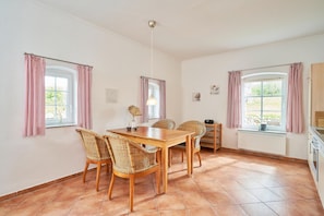 Un coin repas avec une table en bois et des chaises en osier, deux fenêtres avec des rideaux, un radiateur et un espace cuisine avec des placards.