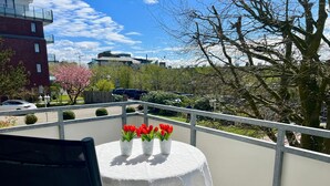 A balcony with a round table covered in a white tablecloth, three white vases with red flowers, and a view of a parking lot and buildings in the background. - N 6 (BC.a5) - Stranddomicil Steiner (Büsum)
