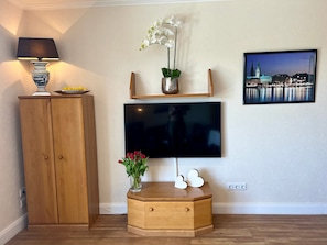 Ein Wohnzimmer mit an der Wand montiertem Fernseher, einem Holzschrank, einem kleinen Tisch mit Lampe, einer Vase mit Blumen und einem Bilderrahmen mit Stadtansicht.