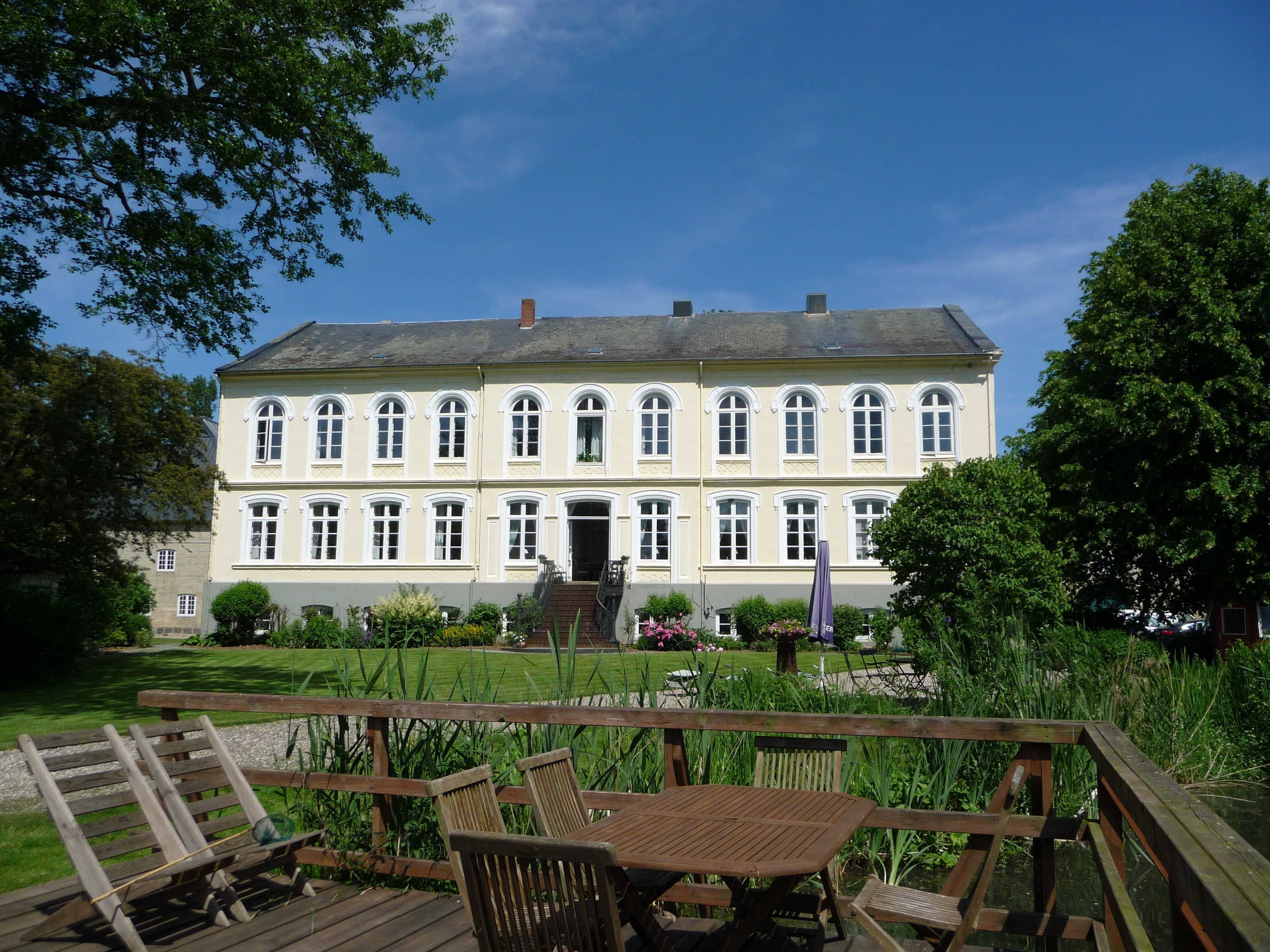 Ein großes, zweistöckiges Gebäude mit symmetrischer Fassade, einem zentralen Eingang und einem umlaufenden Garten mit einer Holzterrasse und Stühlen.