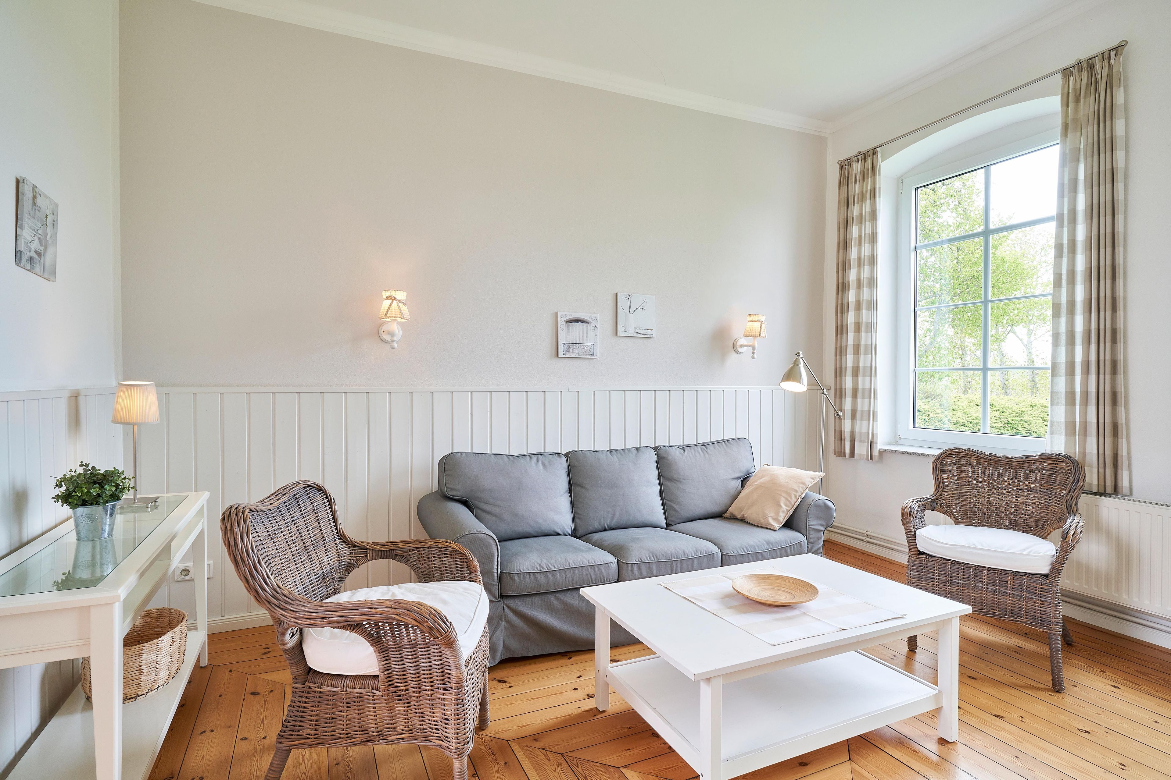 Un salon avec un canapé gris, deux fauteuils en osier, une table basse blanche et une fenêtre avec des rideaux.