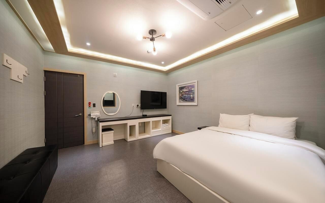 A hotel room with a bed, a desk with a flat-screen TV, a mirror, a door, and a ceiling light fixture.