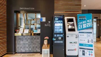 A hotel reception area with a self-check-in kiosk, a digital information board, and a sign promoting QR code check-in.