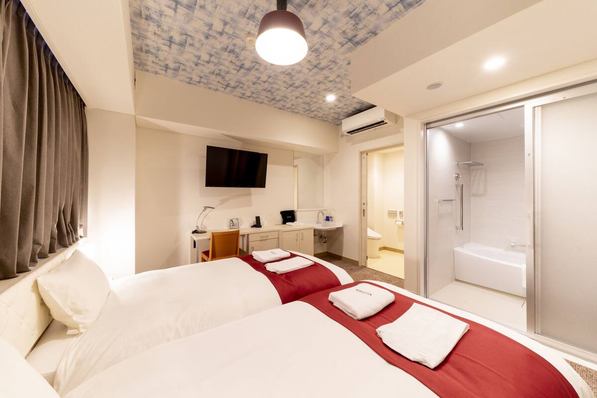 A hotel room with two beds, a desk, a TV, a bathroom with a shower and bathtub, and a ceiling with a textured pattern.