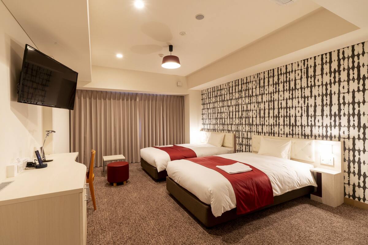 A hotel room with two beds, a desk, a TV, and a patterned wall.