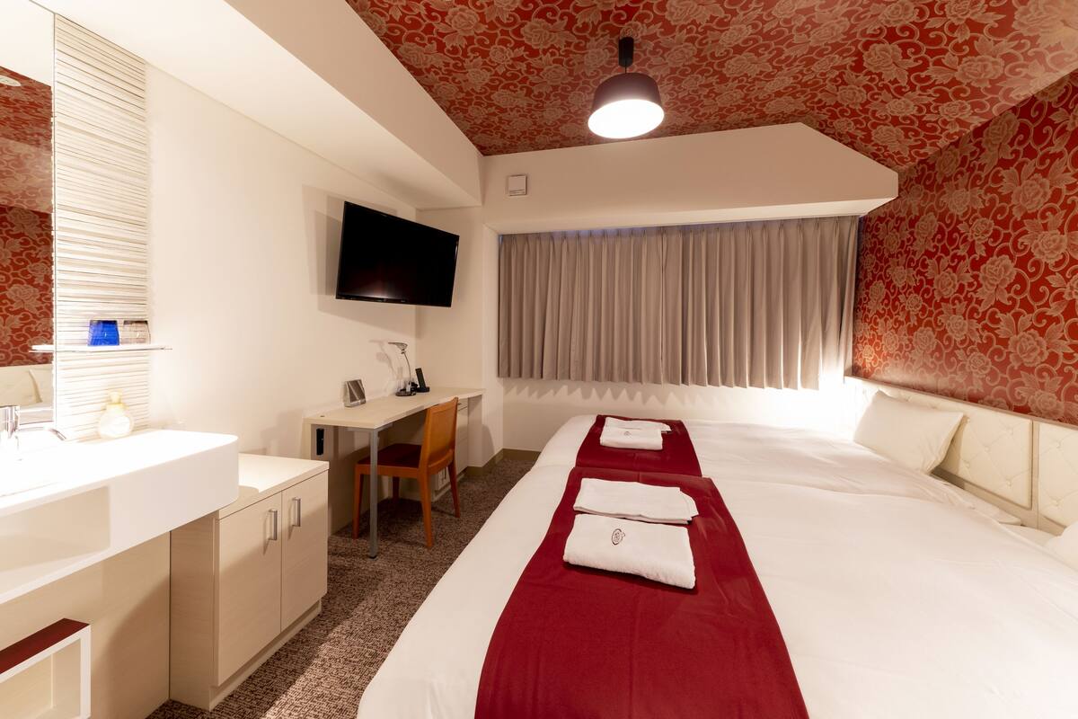A hotel room with a large bed, a desk, a chair, a TV mounted on the wall, and a window with curtains.