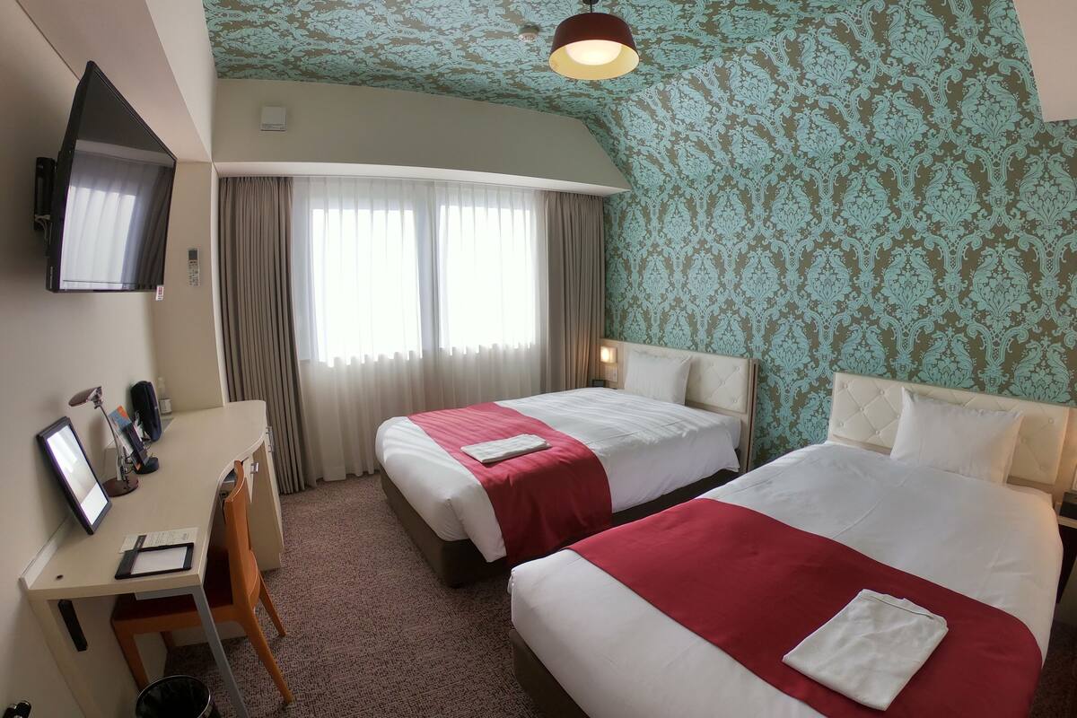 A hotel room with two beds, a desk with a computer, a TV, and a patterned wall.