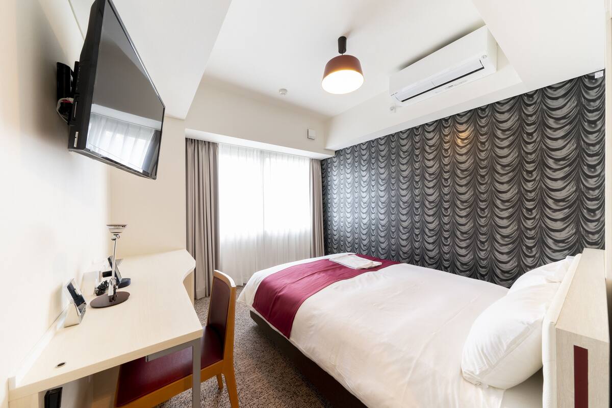 A hotel room with a bed, a desk, a chair, a TV mounted on the wall, and a window with curtains.