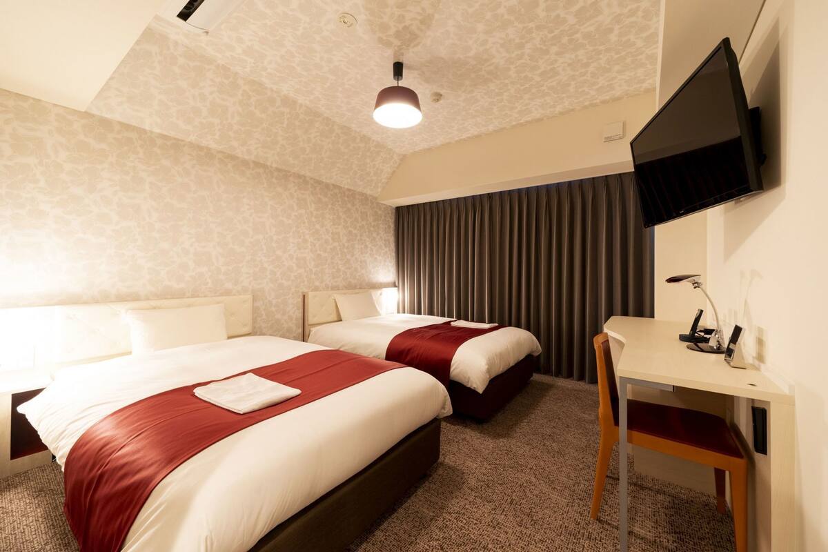 A hotel room with two beds, a desk, a flat-screen TV, and a ceiling light.