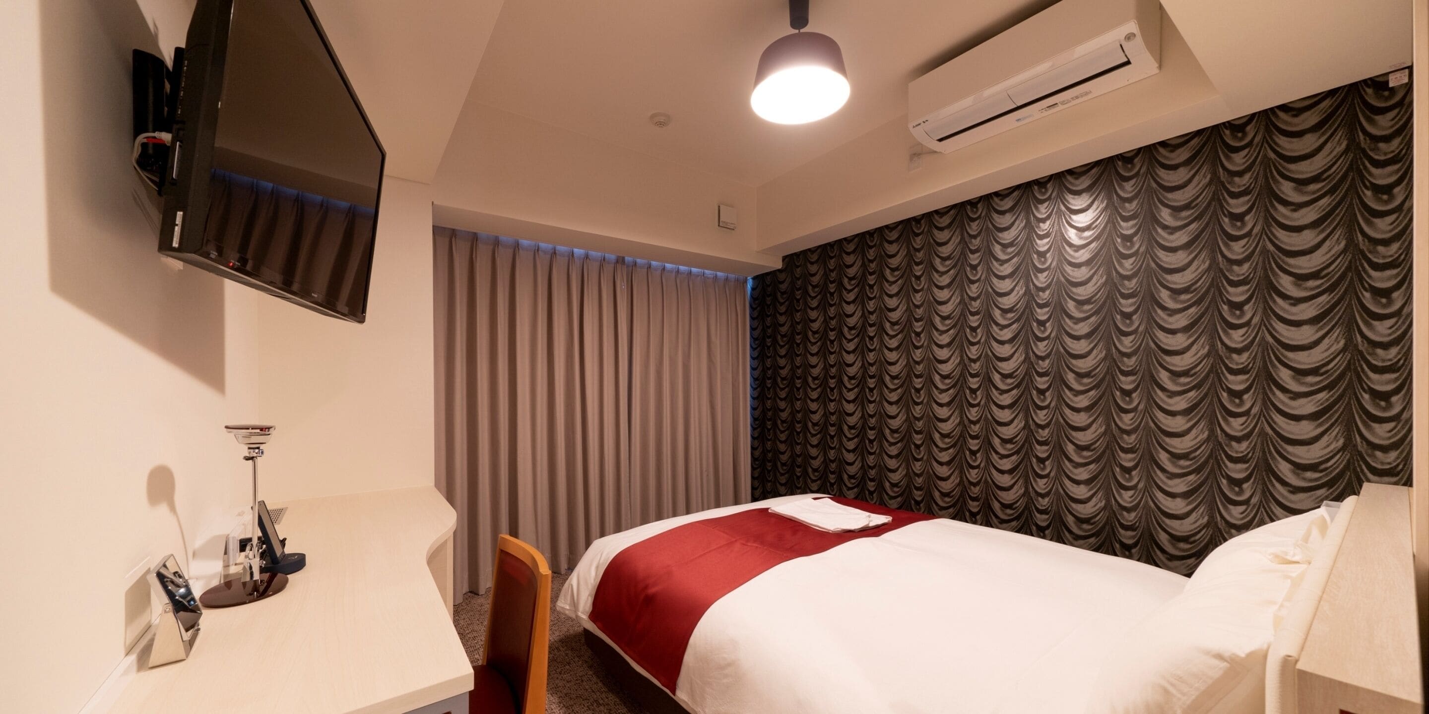 A hotel room with a bed, a desk with a chair, a TV mounted on the wall, and an air conditioning unit.