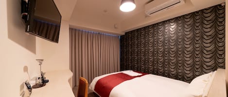 A hotel room with a bed, a desk with a chair, a TV mounted on the wall, and an air conditioning unit.