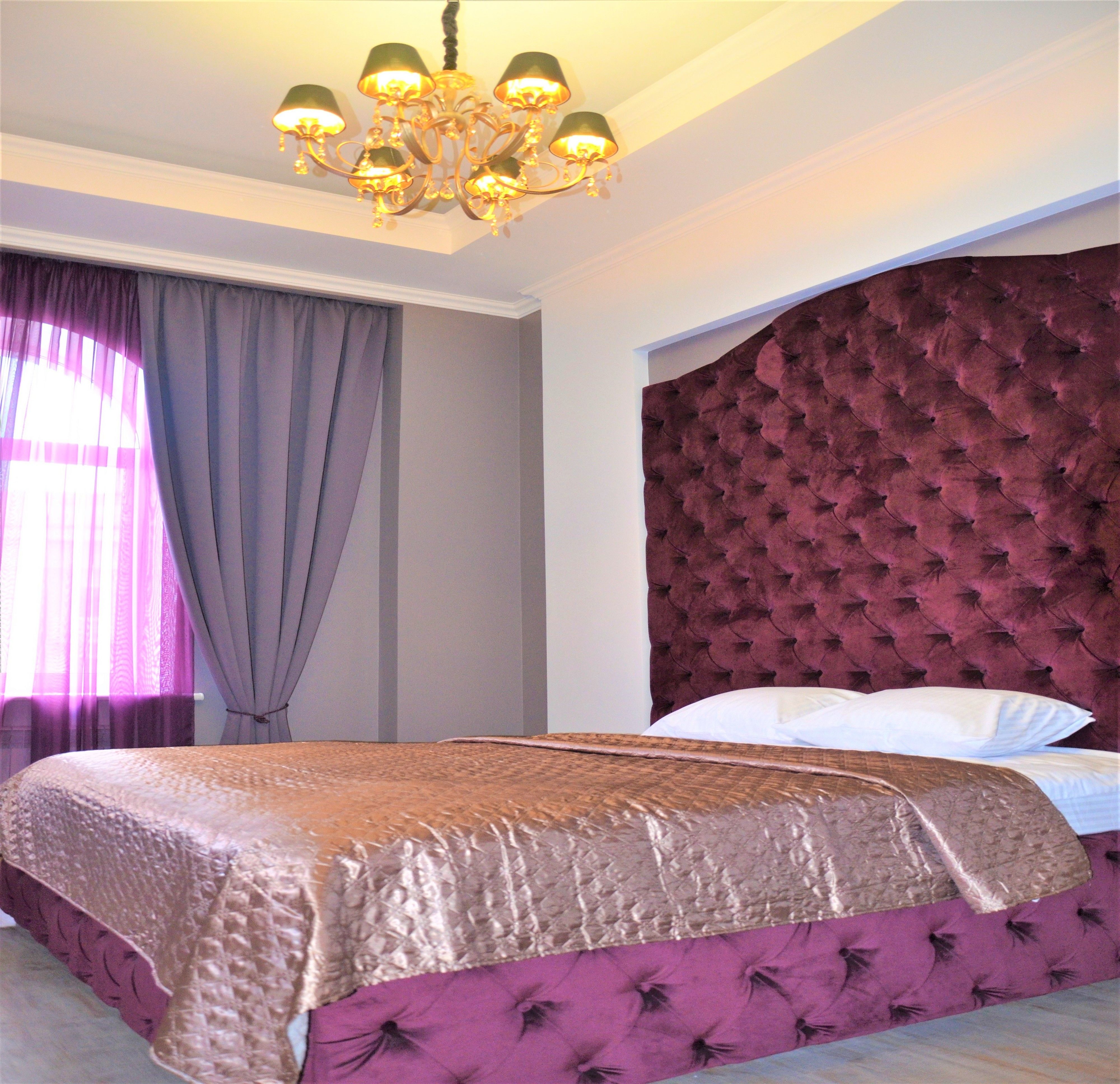 Een hotelkamer met een groot bed, een kroonluchter, een gestoffeerd hoofdeinde en een raam met gordijnen.