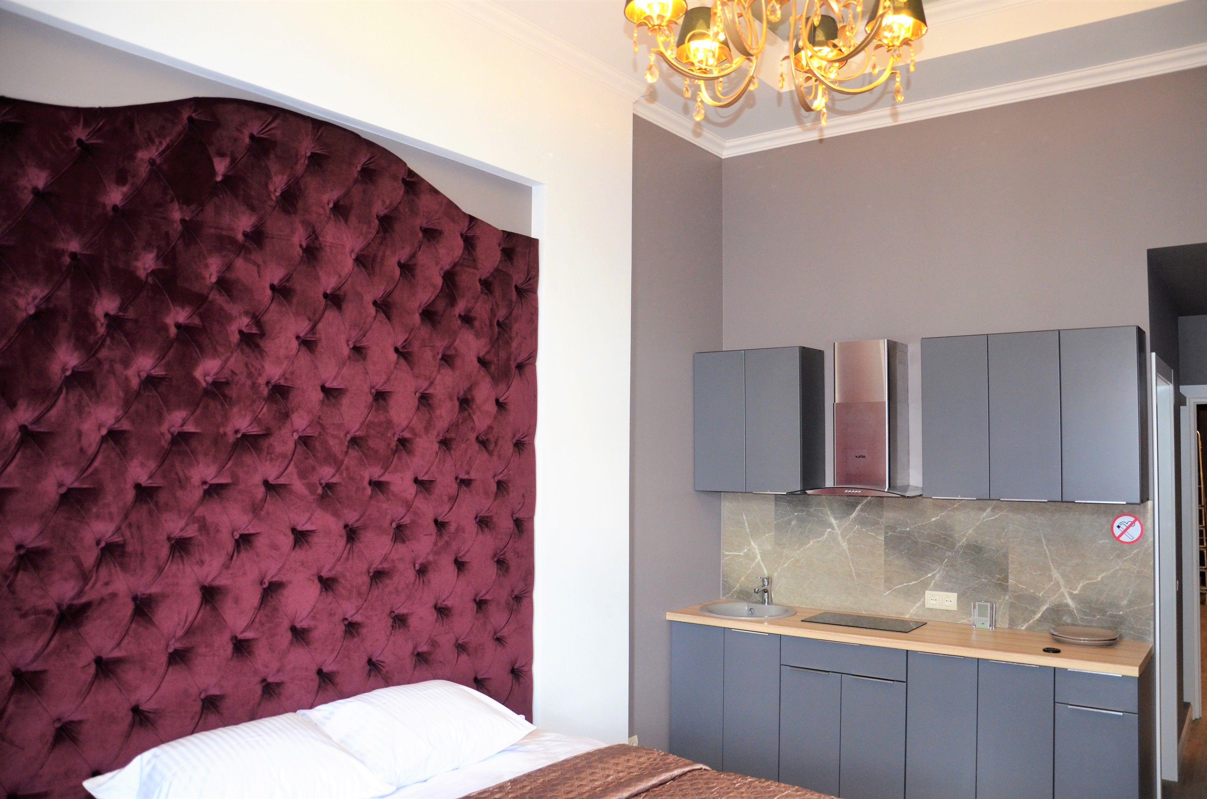 A bedroom with a tufted headboard, a bed, a bedside table, and a built-in kitchen area with cabinets and a countertop.
