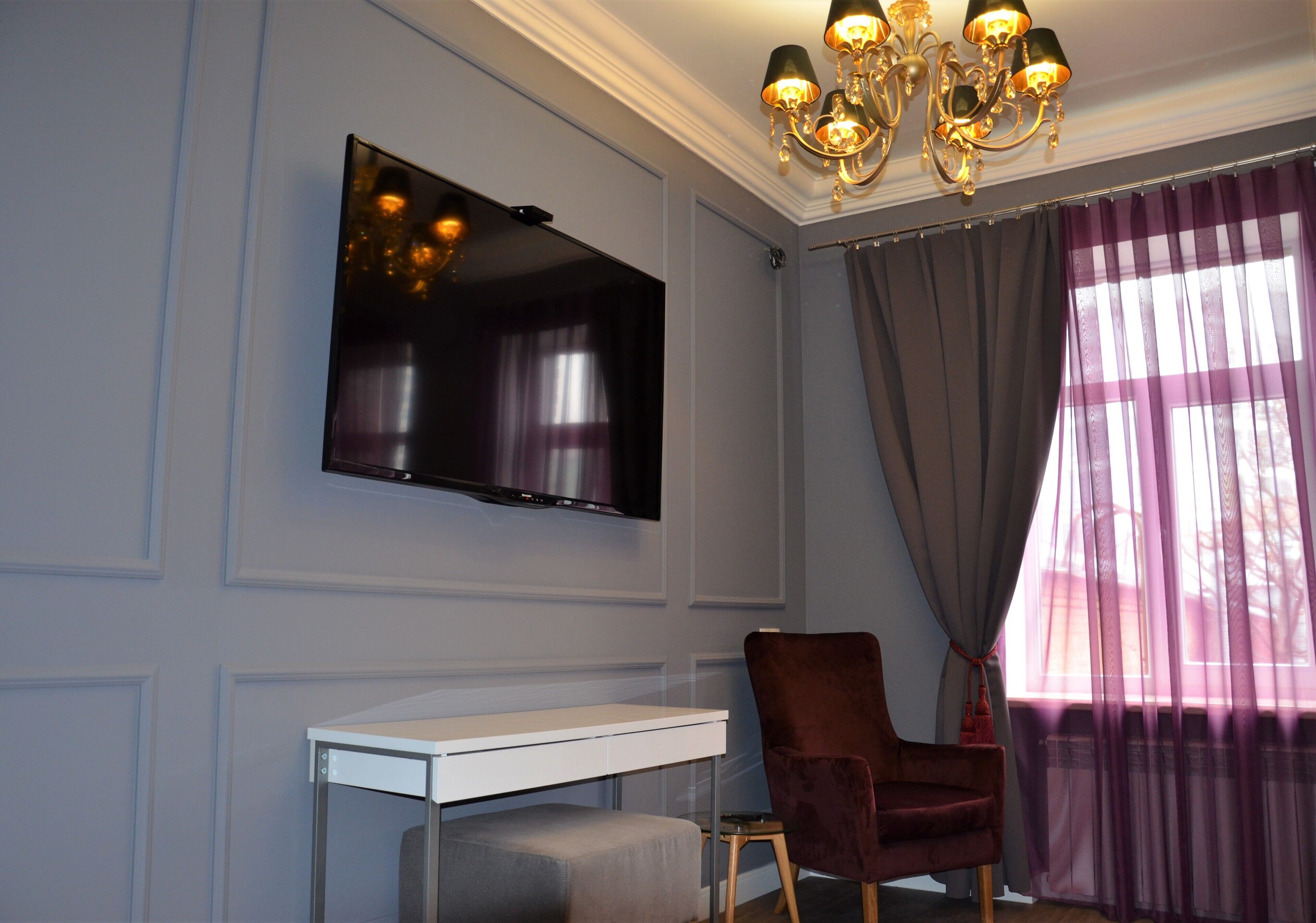 A modern room with a flat-screen TV mounted on the wall, a white desk, a red chair, and a large window with sheer curtains.