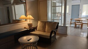 Un coin salon confortable avec un fauteuil moelleux, une petite table ronde sur laquelle se trouve une télécommande et une lampe. Un miroir reflète une partie de la pièce, et une porte donne accès à un autre espace.