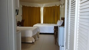 Une chambre d’hôtel avec un lit, un bureau et une chaise. Il y a une fenêtre avec des rideaux, et une porte qui donne sur une autre pièce.