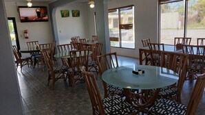 Un espace repas avec des tables rondes et des chaises en bois, un téléviseur fixé au mur et de grandes fenêtres équipées de stores.