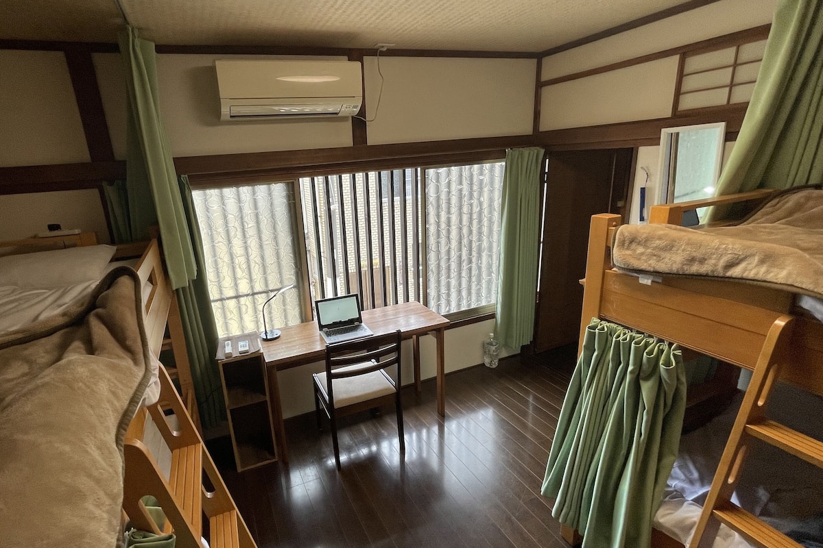 Een kleine kamer met stapelbedden, een bureau met een laptop, een raam met gordijnen en een airconditioning.