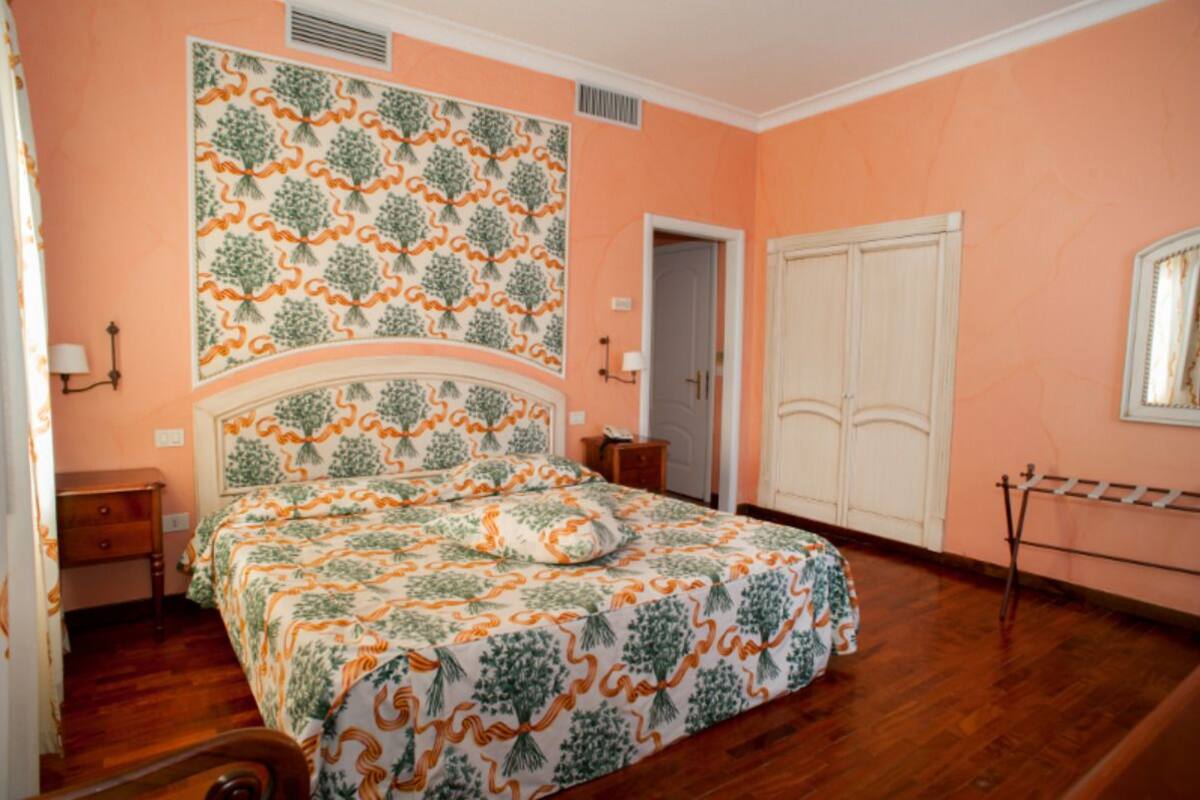 A bedroom with a patterned bedspread, a wooden floor, a wardrobe, and a door leading to another room.