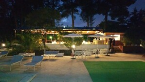 Un espacio exterior con sillones de lounge y sombrillas, una zona de bar con mesas y sillas, y césped artificial verde.