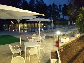 Un área de estar al aire libre con mesas y sillas bajo sombrillas, una piscina y cabañas de madera al fondo.