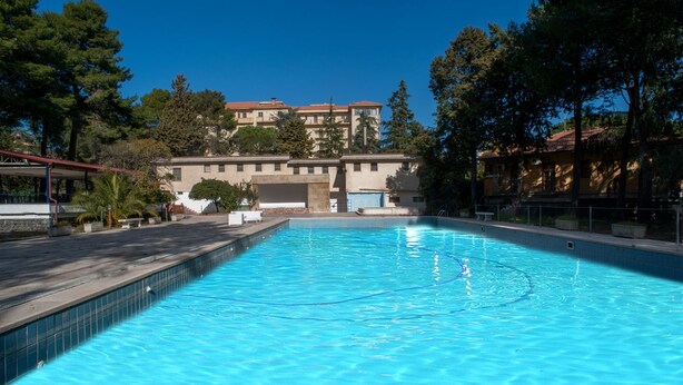 Una piscina rectangular con agua azul cristalina, rodeada de árboles y un edificio con techo rojo al fondo.