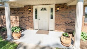 Une maison en briques avec une porte blanche, deux plantes en pot de chaque côté et un porche abrité.