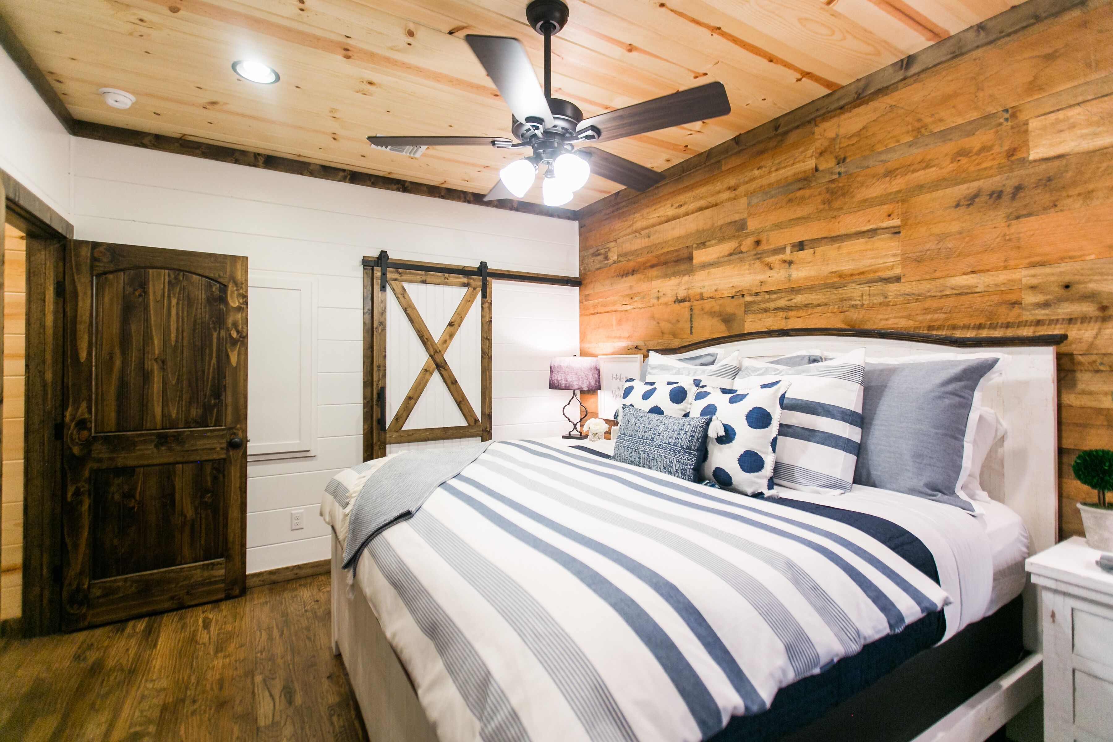Un dormitorio con techo de madera, una cama con ropa de cama a rayas, una mesita de noche con una lámpara y una puerta de madera.