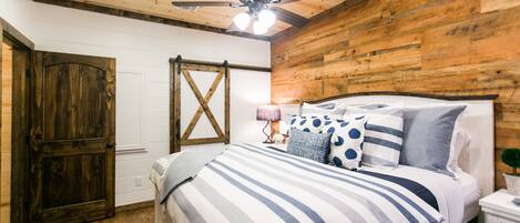 Un dormitorio con techo de madera, una cama con ropa de cama a rayas, una mesita de noche con una lámpara y una puerta de madera.