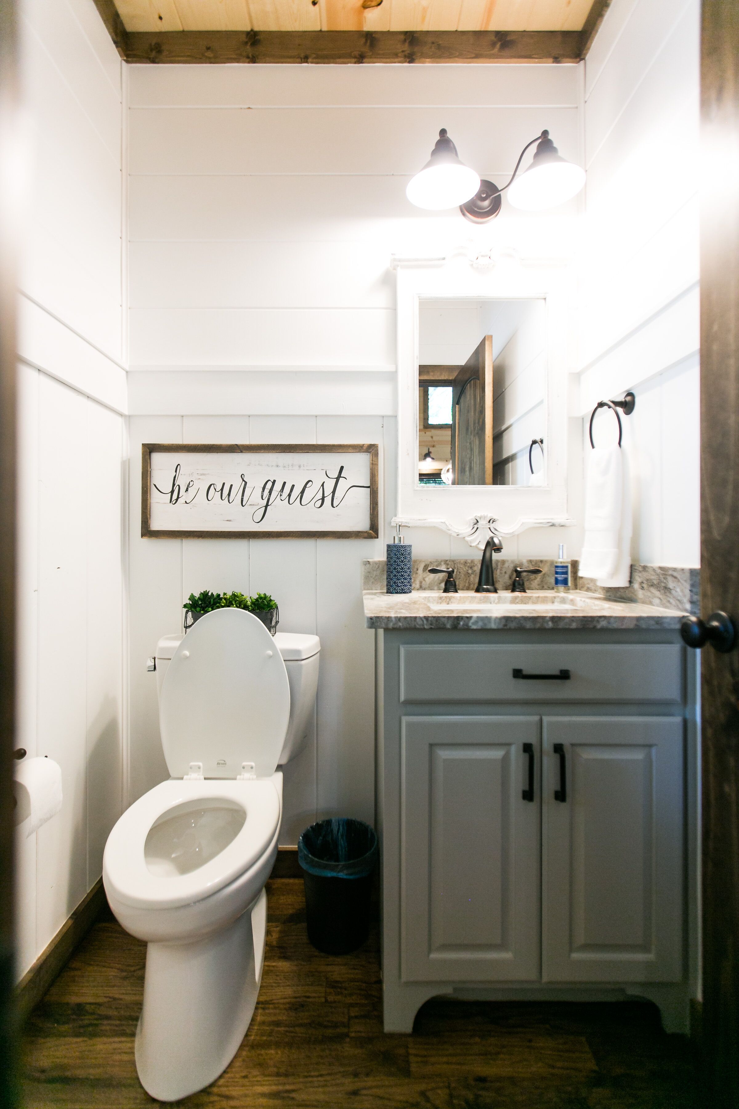A bathroom with a white toilet, a sink with a mirror above it, and a sign that says "be our guest".