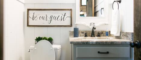 A bathroom with a white toilet, a sink with a mirror above it, and a sign that says "be our guest".