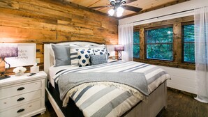 A bedroom with a wooden ceiling, a bed with striped bedding, a nightstand with a lamp, and a window with blinds. - Free at Last- Romantic, Hot Tub, Loft, Free Firewood, Great Location! (Broken Bow)