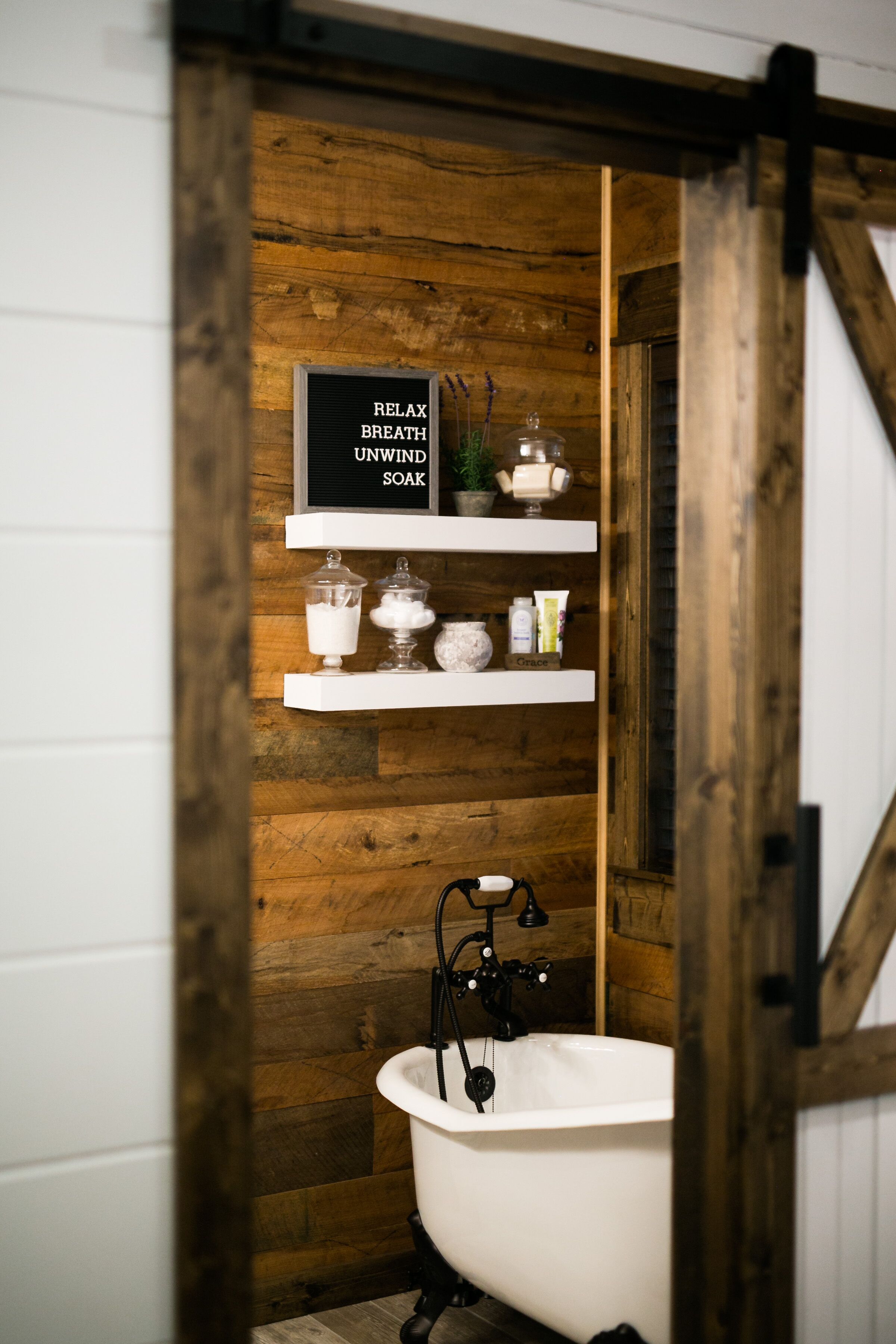 A rustic bathroom with a wooden wall, a white bathtub, and shelves with toiletries.