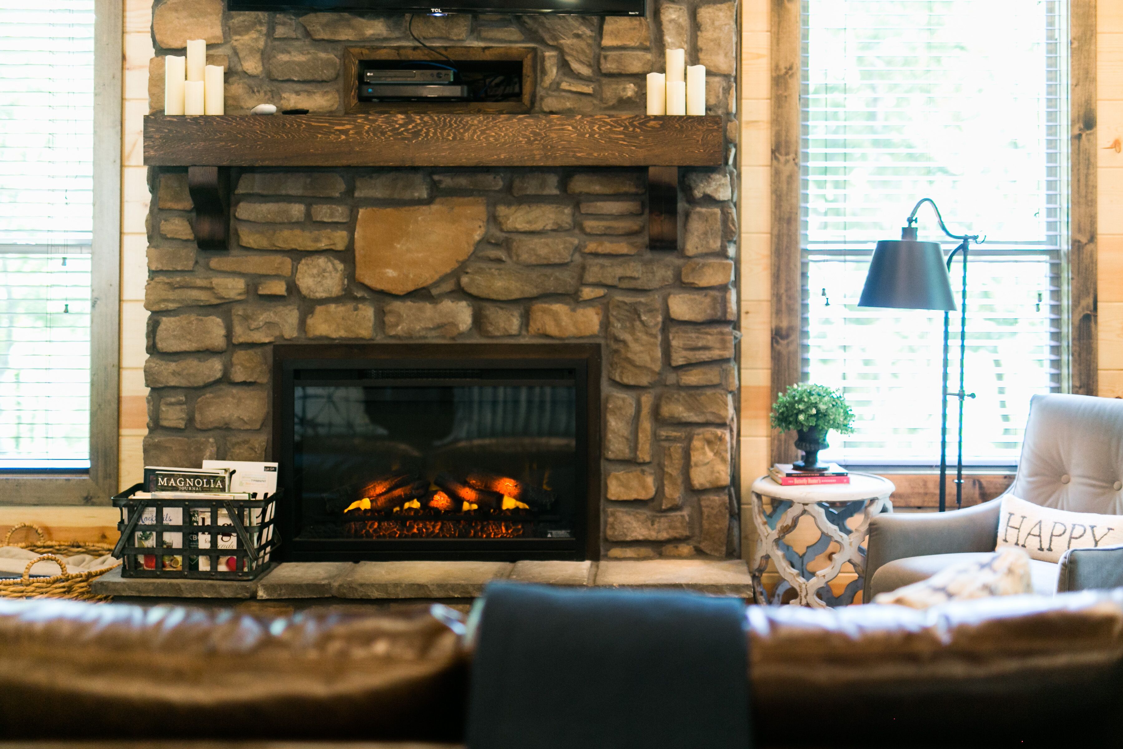 A stone fireplace with a fire, a cozy seating area with a 'HAPPY' pillow, and a small side table with a lamp.