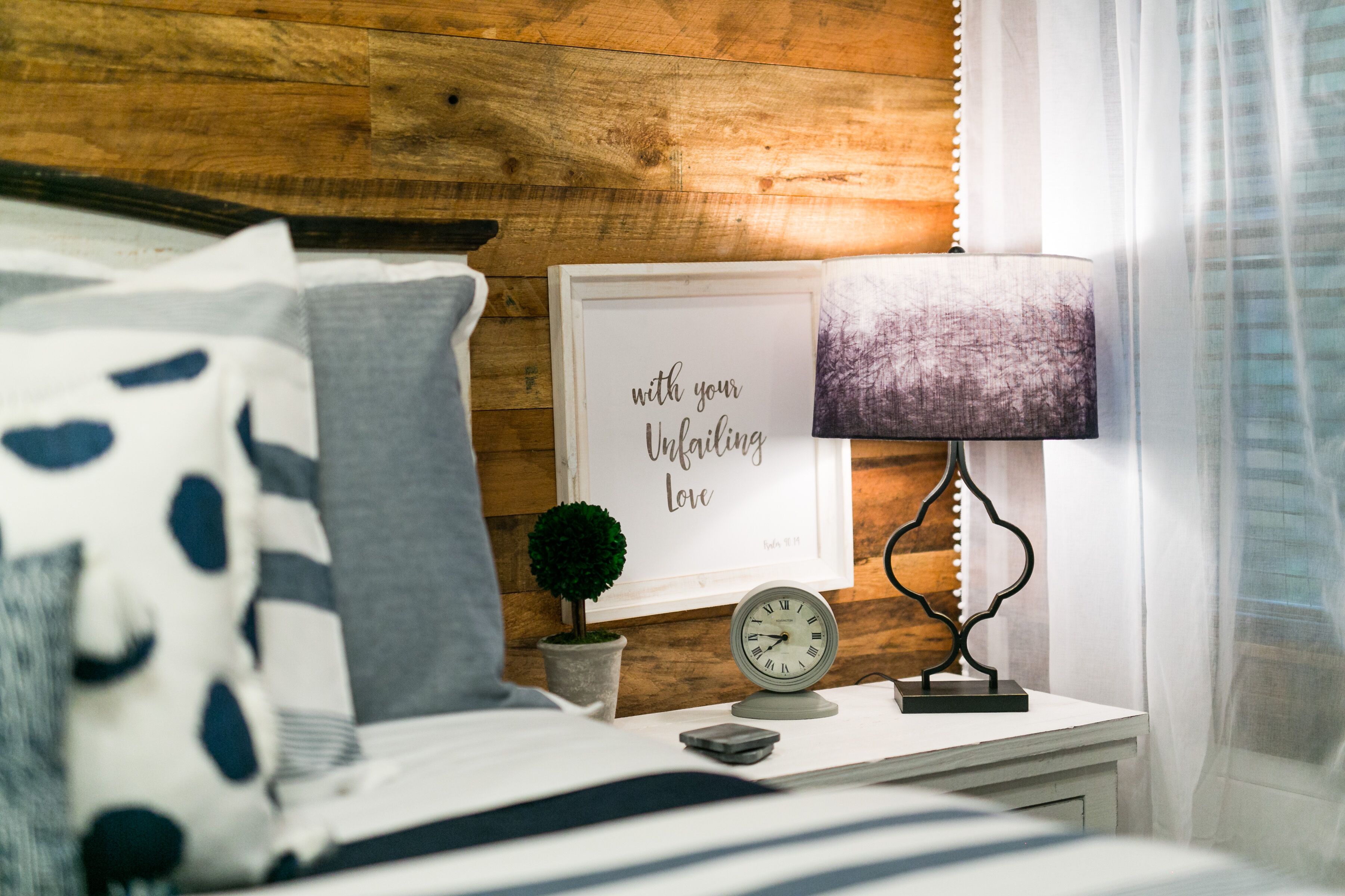 Un dormitorio ordenado con cama, mesita de noche con reloj, cita enmarcada, lámpara y una pequeña planta en maceta.