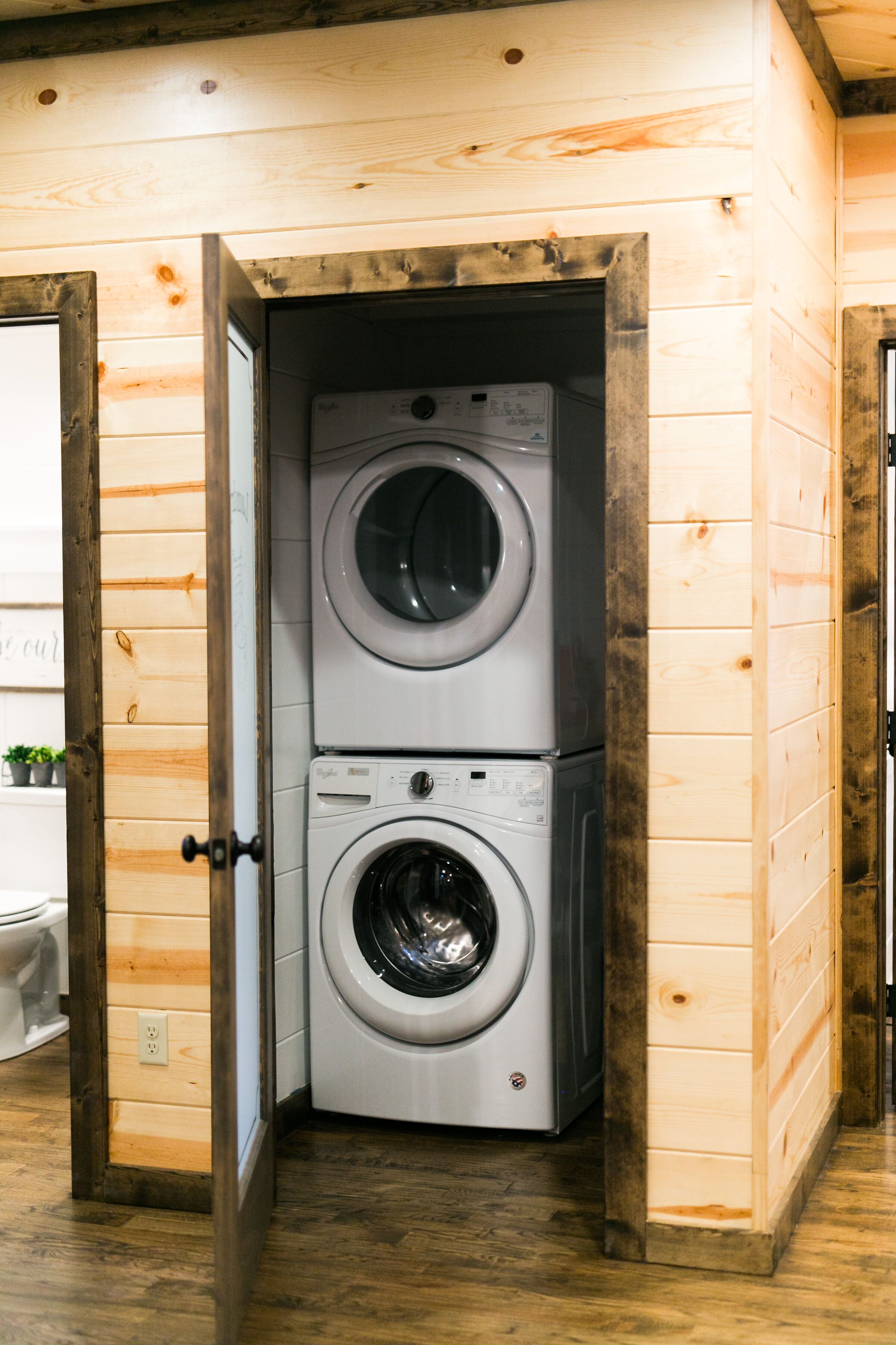 A laundry room with two stacked washing machines, wooden walls, and a door leading to another room.