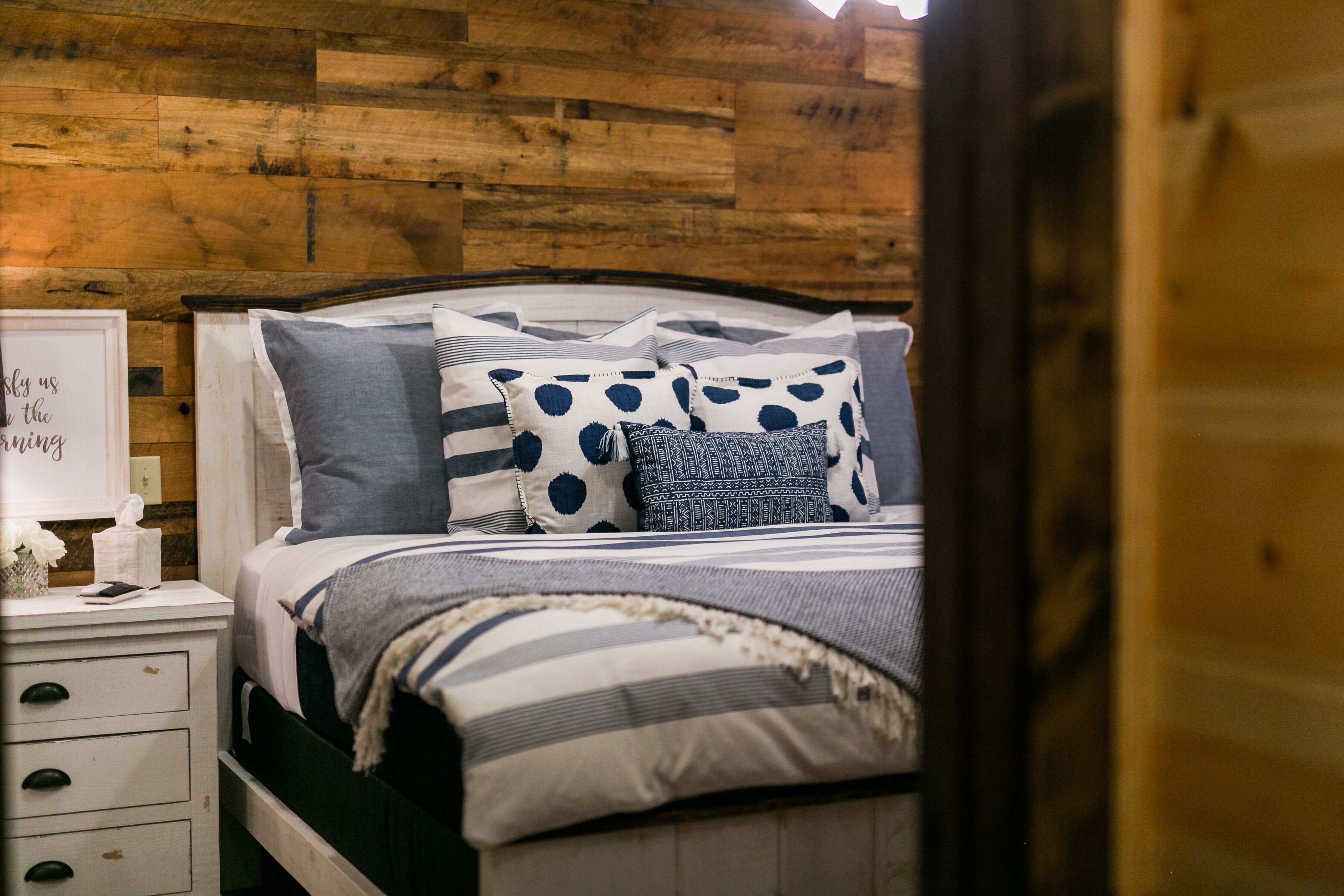A neatly made bed with striped bedding and polka dot pillows, a bedside table with a lamp, and a wooden wall.