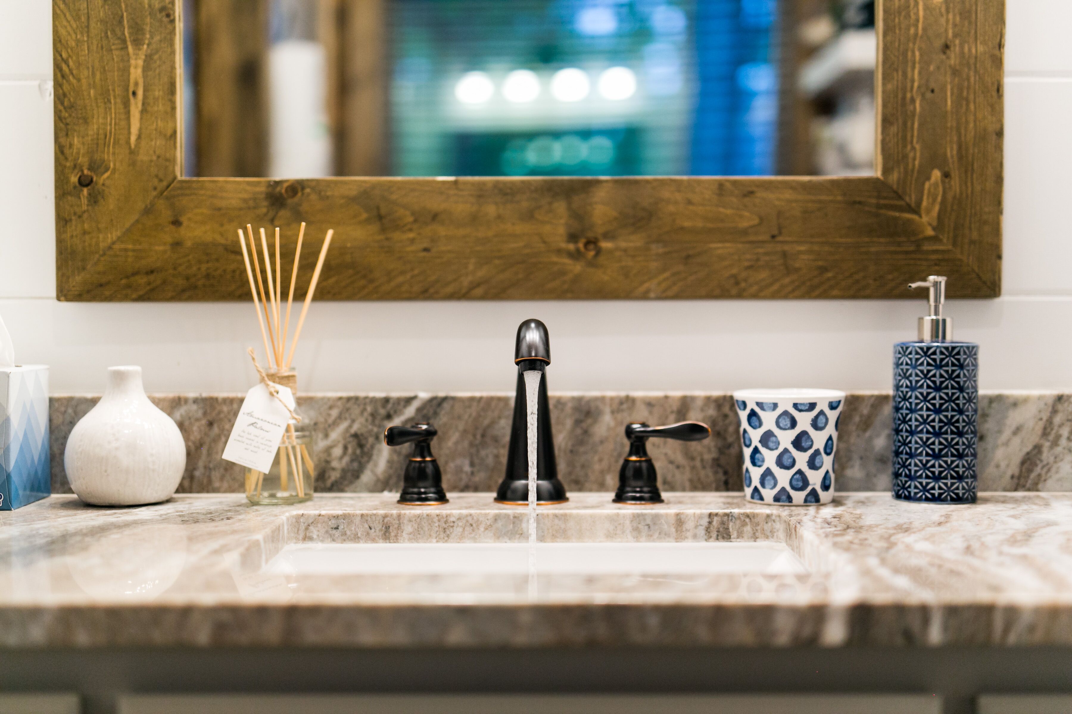 A bathroom sink with a wooden-framed mirror above it, a marble countertop, and various toiletries including a white vase, reed diffuser, and patterned containers.