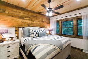 A bedroom with a wooden ceiling, a bed with striped bedding, a nightstand with a lamp, and a window with blinds. - Free at Last- Romantic, Hot Tub, Loft, Free Firewood, Great Location! (Broken Bow)